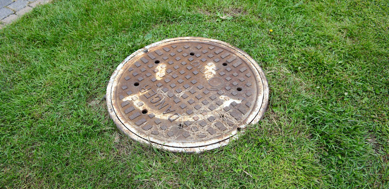 An image of A photograph of a fairly rusted manhole cover with a white smiley face spray-painted on it surrounded by grass and a brick path to the upper left hand corner of the frame